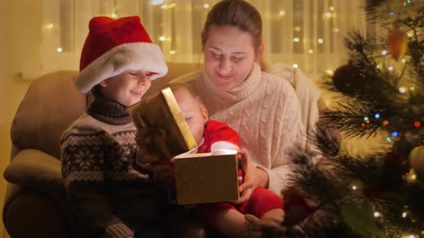 Schattige baby jongen zittend op moeders schoot in een fauteuil en kijkend in gloeiende kerstdoos. Gezinnen en kinderen vieren wintervakantie. — Stockvideo