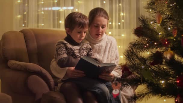 Leuke jongen met moeder zit fauteuil naast de kerstboom en het lezen van groot boek. Gezinnen en kinderen vieren wintervakantie. — Stockvideo