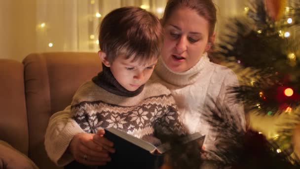 Glad leende pojke med mor läser sagobok bredvid glödande dekorerad julgran. Familjer och barn som firar vinterlov. — Stockvideo