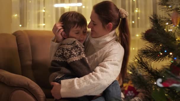 Sonriente madre joven abrazando a su hijo sentado en el sillón junto al árbol de Navidad en la sala de estar. Familias y niños celebrando las vacaciones de invierno. — Vídeo de stock