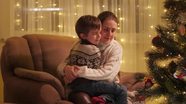 Gelukkige moeder en zoon zitten in een fauteuil naast de kerstboom en knuffelen. Gezinnen en kinderen vieren wintervakantie. — Stockvideo
