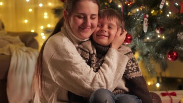Portret van een gelukkige moeder die haar zoon knuffelt en kust op kerstavond naast een stralende kerstboom. Gezinnen en kinderen vieren wintervakantie. — Stockvideo