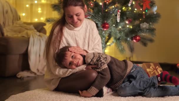 Junge Mutter streichelt ihren lächelnden Sohn, der neben dem leuchtenden Weihnachtsbaum liegt. Familien und Kinder feiern Winterurlaub. — Stockvideo