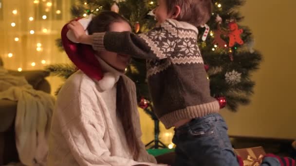 Menino sorridente feliz se divertindo e colocando chapéu de Papai Noel na mãe ao lado da árvore de Natal — Vídeo de Stock