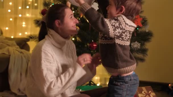 Noel arifesinde annesine Noel şapkası takan süveter giyen gülen çocuk. Aileler ve çocuklar kış tatilini kutluyor. — Stok video