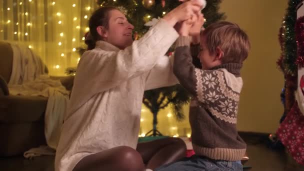 Genç anne oğluna Noel Baba şapkası takıyor ve Noel ağacının altında ona sarılıyor. Aileler ve çocuklar kış tatilini kutluyor. — Stok video