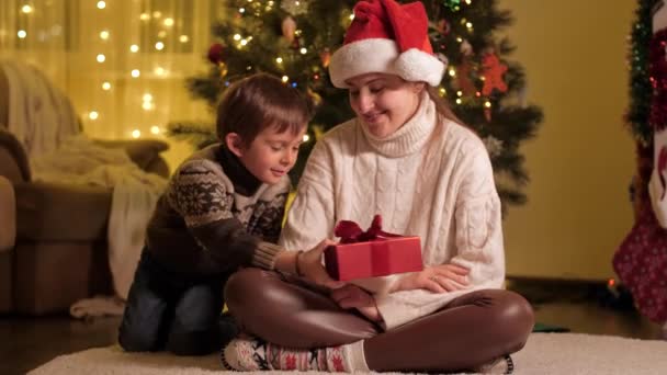 Alegre niño sonriente con la madre dando regalos de Navidad y abrazándose bajo el árbol de Navidad en casa. Familias y niños celebrando las vacaciones de invierno. — Vídeo de stock