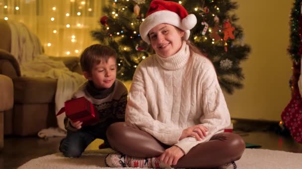 Niño haciendo sorpresa navideña y regalando a su madre con sombrero de Santa Claus. Familias y niños celebrando las vacaciones de invierno. — Vídeos de Stock