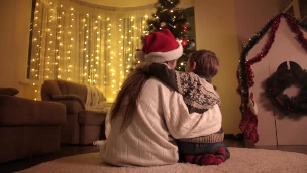 Genç anne küçük oğluna sarılıyor ve oturma odasında parlayan Noel ağacına bakıyor. Aileler ve çocuklar kış tatilini kutluyor. — Stok video