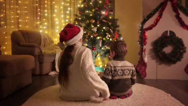 Mutlu bir ailenin yerde oturup renkli Noel ağaçlarının ışıklarına baktığı arka manzara. Aileler ve çocuklar kış tatilini kutluyor. — Stok video