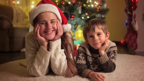 Mosolygós fiú anyával a szőnyegen fekszik a karácsonyfa alatt, miközben ünnepli az újévet. A téli ünnepeket ünneplő családok és gyermekek. — Stock videók