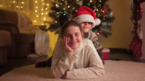 喜庆的男孩躺在母亲身旁,旁边点缀着闪闪发光的圣诞树.庆祝寒假的家庭和儿童. — 图库视频影像