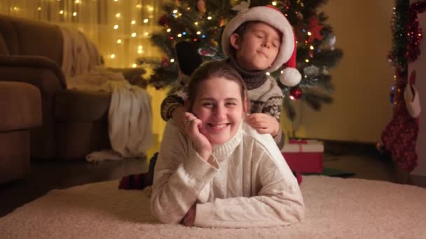 Netter Junge mit Mutter entspannt auf dem Boden und umarmt neben dem Weihnachtsbaum. Pure Emotionen bei Familien und Kindern, die Winterurlaub feiern. — Stockvideo