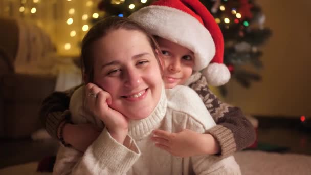 Portrait rapproché du petit garçon au chapeau de Père Noël embrassant sa mère à côté du sapin de Noël. Pures émotions des familles et des enfants qui célèbrent les vacances d'hiver. — Video