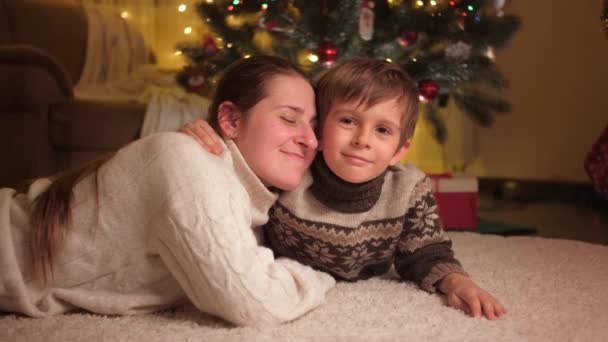 Portrait of smiling mom with son embracing and lying on carpet under Christmas tree in living room. Pure emotions of families and children celebrating winter holidays. — Stock Video