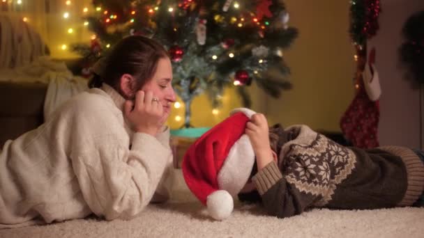 Lachende jongen met moeder ligt oog in oog op tapijt onder de kerstboom en verbleekt met de kerstmuts. Pure emoties van gezinnen en kinderen die de wintervakantie vieren. — Stockvideo