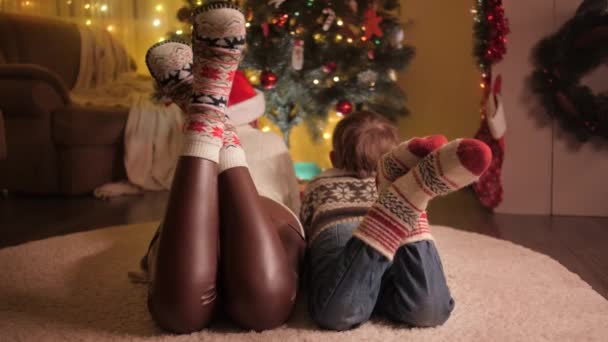 Familia con calcetines de lana de punto relajante en la alfombra y mirando en el árbol de Navidad decorado con luces de guirnalda de colores. Las emociones puras de las familias y los niños que celebran las vacaciones de invierno. — Vídeo de stock