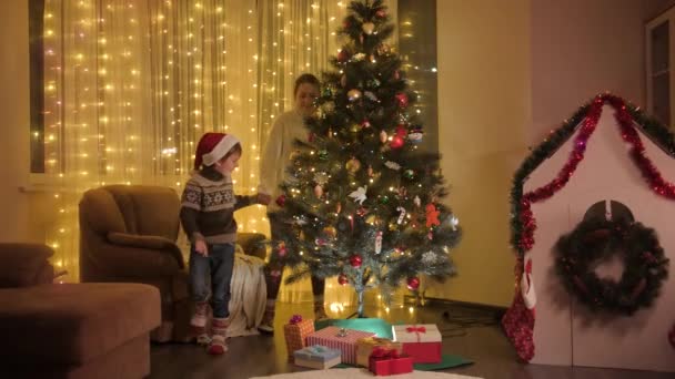 快乐的母亲带着儿子在客厅里跳着欢快的圣诞树。庆祝寒假的家庭和孩子的纯真感情. — 图库视频影像