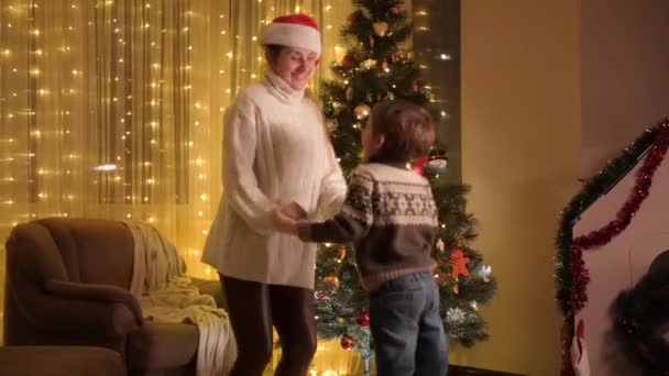 Neşeli çocuk, annesi dans ediyor ve parıldayan Noel ağacının yanında zıplıyor. Aile ve çocukların kış tatilini kutladıkları saf duygular.. — Stok video