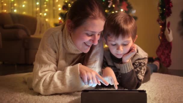 Ritratto di mamma sorridente con figlio che naviga su internet e guarda video su tablet sotto l'albero di Natale. Emozioni pure di famiglie e bambini che festeggiano vacanze invernali. — Video Stock