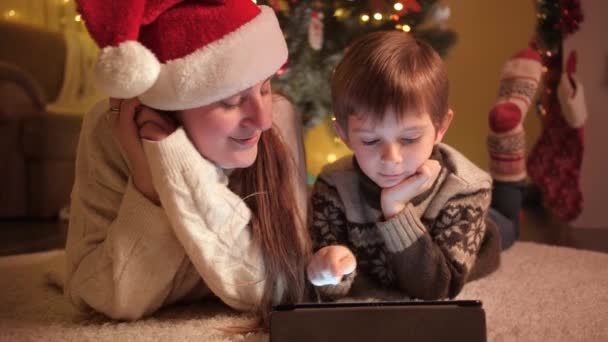 Anya gyermekkel karácsony este digitális táblagéppel a karácsonyfa mellett. A téli ünnepeket ünneplő családok és gyermekek tiszta érzelmei. — Stock videók