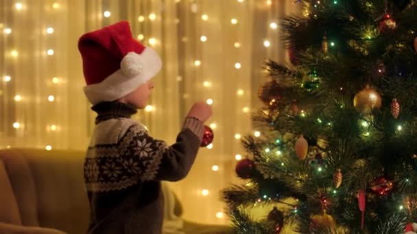 Noel Baba şapkalı yakışıklı çocuk yeni yıl kutlamaları için Noel ağacını süslüyor. Aile ve çocukların kış tatilini kutladıkları saf duygular.. — Stok video