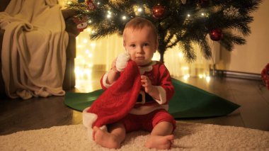 Noel Baba kostümlü şirin, gülümseyen bir çocuk oturma odasında süslü Noel ağacının altında oturuyor. Aileler ve çocuklar kış tatilini kutluyor.