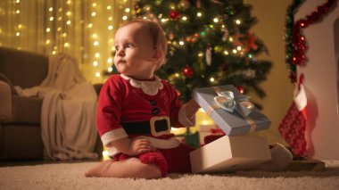 Noel Baba kostümü giymiş, Noel ağacının altında elinde hediye kutusuyla oturan tatlı bir bebek. Aileler ve çocuklar kış tatilini kutluyor.