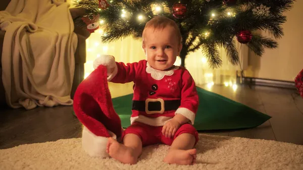 Noel Baba kostümlü şirin bir bebek Noel ağacının altında oturuyor ve kameraya gülümsüyor.