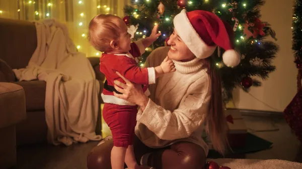 サンタクロースの衣装を着た可愛い男の子がクリスマスツリーの隣に座っています。冬の休日を祝う家族や子供. — ストック写真