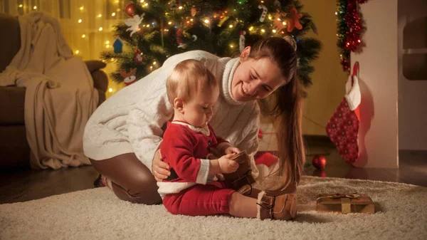 Jovem mãe sorridente vestindo bota quente para seu filho bebê em traje de Papai Noel ao lado da árvore de Natal. Famílias e crianças comemorando as férias de inverno. — Fotografia de Stock