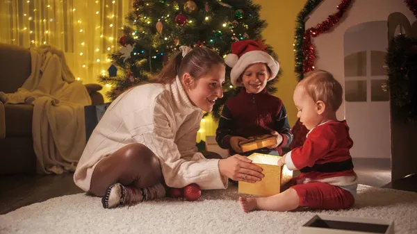 Šťastná matka s dítětem a starší syn představuje dárkové krabice vedle vánočního stromečku v domě. Rodiny a děti slaví zimní prázdniny. — Stock fotografie