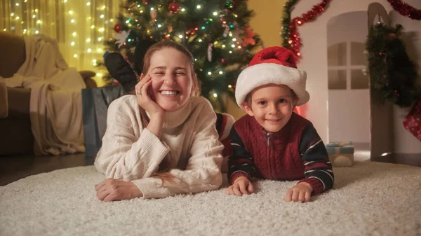 Mãe sorridente com filho deitado no chão ao lado da árvore de Natal e se divertindo. Famílias e crianças comemorando as férias de inverno. — Fotografia de Stock