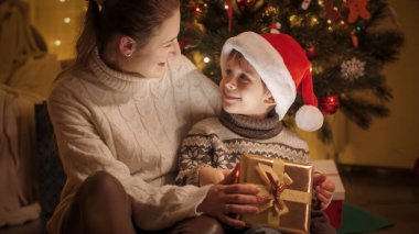 Noel Baba şapkalı gülümseyen çocuğun portresi. Altın hediye kutusundaki Noel hediyesinden sonra annesine gülümsüyor.