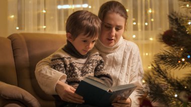 Genç anne oğluna kitap okuyor. Parlayan Noel ağacının yanındaki koltukta oturuyor. Aileler ve çocuklar kış tatilini kutluyor.