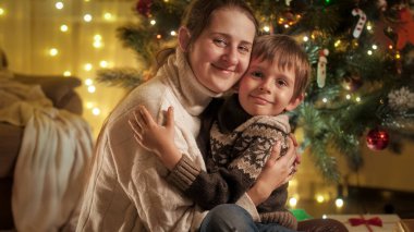 Annesiyle Noel ağacının yanında sarılıp kameraya bakan neşeli gülümseyen çocuğun portresi. Aileler ve çocuklar kış tatilini kutluyor.