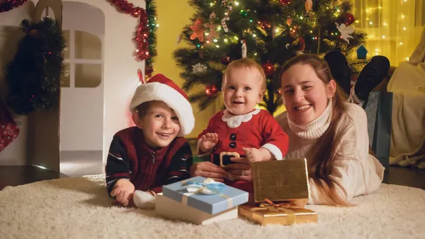 Mère joyeuse avec ses enfants couchés sur le sol sous l'arbre de Noël lumineux dans la maison. Familles et enfants célébrant les vacances d'hiver. — Photo