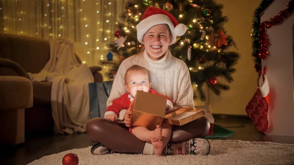 Noel hediyesini tutan mutlu bir anne ve Noel ağacının altında oturan küçük bir çocuk. — Stok fotoğraf