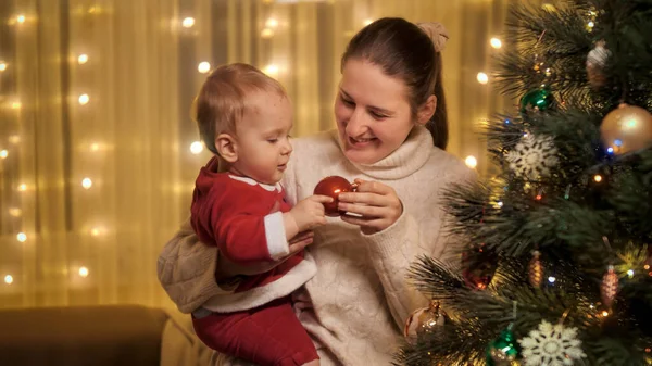 Feliz madre sonriente sosteniendo pequeño hijo bebé y mirando el árbol de Navidad brillante decorado. Familias y niños celebrando las vacaciones de invierno. —  Fotos de Stock