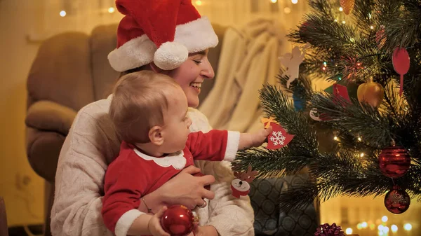 Pequeño niño buscando en el brillante árbol de Navidad y quitándose las bolas de Navidad. Familias y niños celebrando las vacaciones de invierno. — Foto de Stock