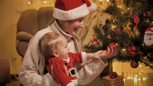 Retrato de jovem mãe mostrando bugigangas em decorações na árvore de Natal para seu filho bebê. Famílias e crianças comemorando as férias de inverno. — Fotografia de Stock