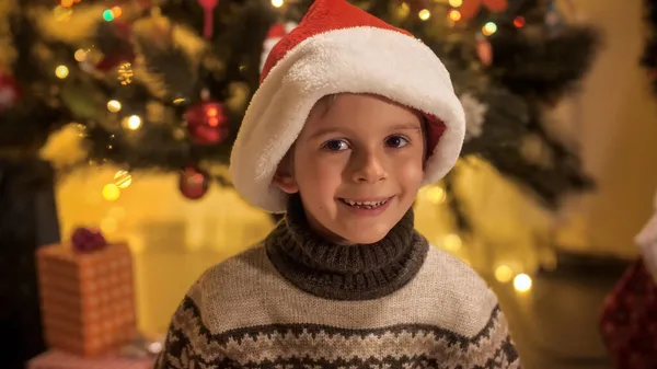 Retrato de menino em chapéu de Papai Noel ao lado da árvore de Natal sorrindo na câmera. Famílias e crianças comemorando as férias de inverno. — Fotografia de Stock
