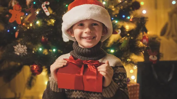 Retrato de niño sonriente alegre con caja de regalo de Navidad con sombrero de Santa sentado al lado del brillante árbol de Navidad decorado. Familias y niños celebrando las vacaciones de invierno. — Foto de Stock