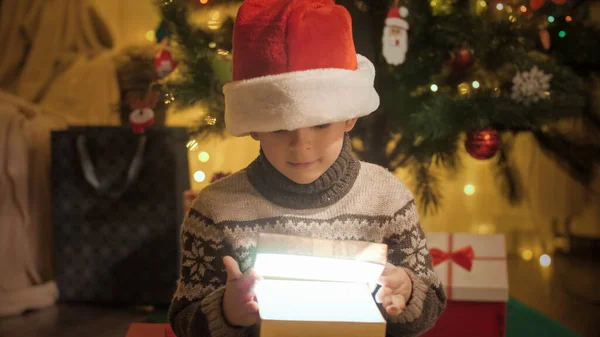 Πορτρέτο του ενθουσιασμένοι αγόρι σε Σάντα καπέλο ανοίγει χριστουγεννιάτικο κουτί δώρο και κοιτάζει μέσα. Χειμερινές διακοπές μαγεία. Οικογένειες και παιδιά γιορτάζουν χειμερινές διακοπές. — Φωτογραφία Αρχείου