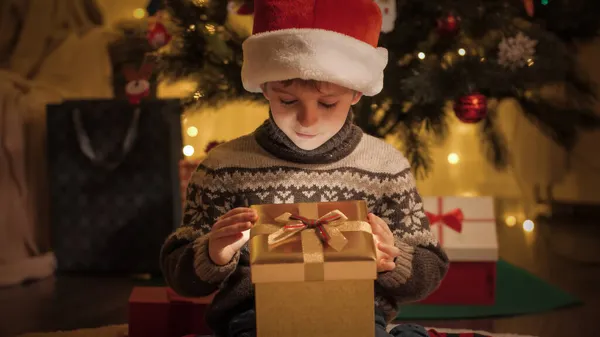 Portret uroczego chłopca w kapeluszu Mikołaja trzymając złote pudełko i patrząc do środka na Boże Narodzenie prezent — Zdjęcie stockowe