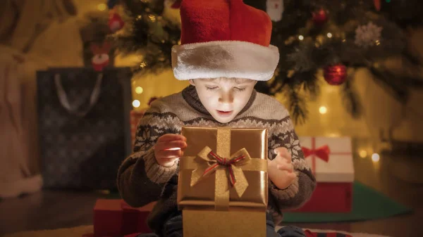 Happy smiling boy in sweater and Santa hat opens Christmas present box and looks inside — Stock Photo, Image