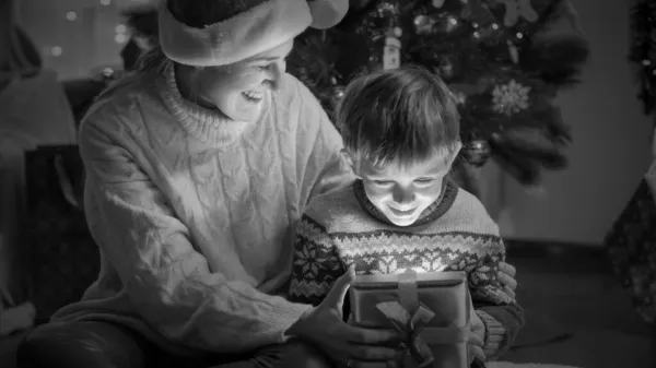 Svart och vitt porträtt av glad liten pojke med mor tittar inuti glödande presentförpackning med julklapp — Stockfoto
