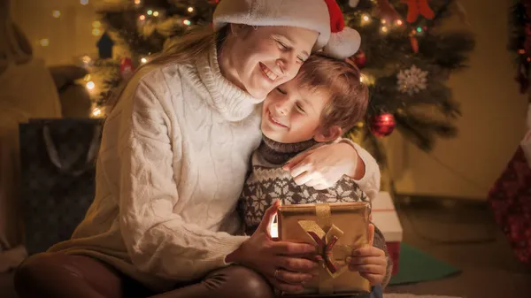 Glad leende mor och son kramas efter att ha fått julklappar under julgranen hemma — Stockfoto