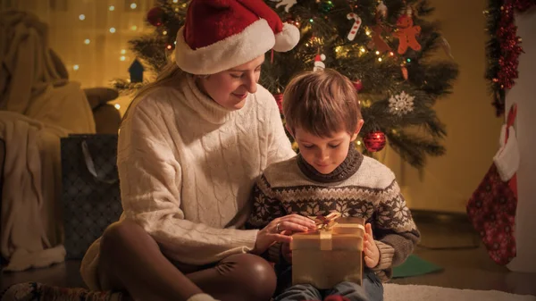 幸せな母親は彼女の息子を抱きかかえてクリスマスプレゼントを包み、リビングルームに存在します。冬の休日を祝う家族や子供. — ストック写真