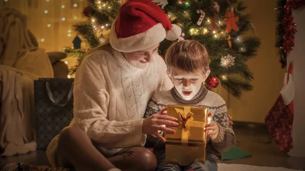 Menino incrível sentado com a mãe sob a árvore de Natal e olhando dentro da caixa de presente de Natal — Fotografia de Stock